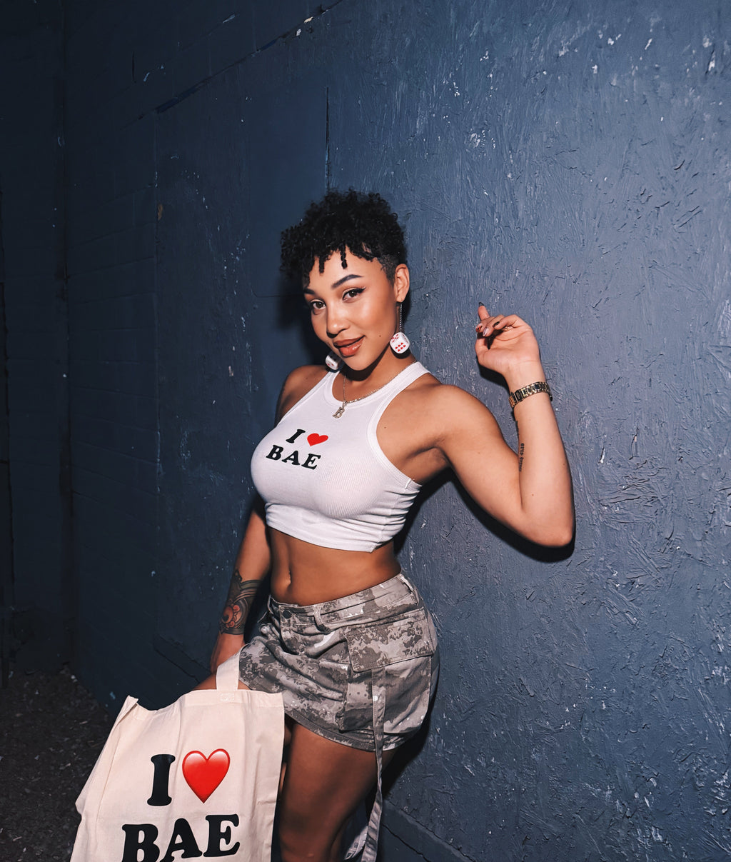 Baelyholding a 'I ❤️ BAE' tote bag against a dark wall.